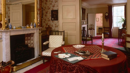 An image of the parlour at Carlyle's House in London showing the interior of a room with an ornate grey marble fireplace surround with a gold gilt framed mirror above, circular wooden table and richly patterned red tablecloth and floor coverings